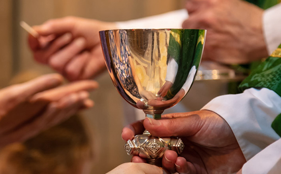 Close-up: Communion chalice (wine-cup) being offered in foreground, Host in background: hands only shown.